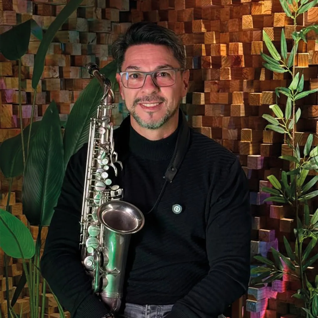 Músico de óculos e barba com saxofone pendurado no pescoço, diante de parede de madeira e plantas