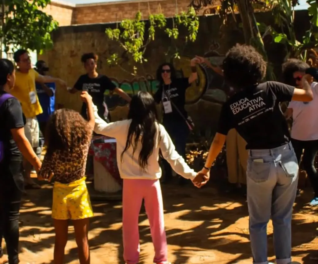 Crianças e educadores de mãos dadas em roda, num quintal de terra com árvores