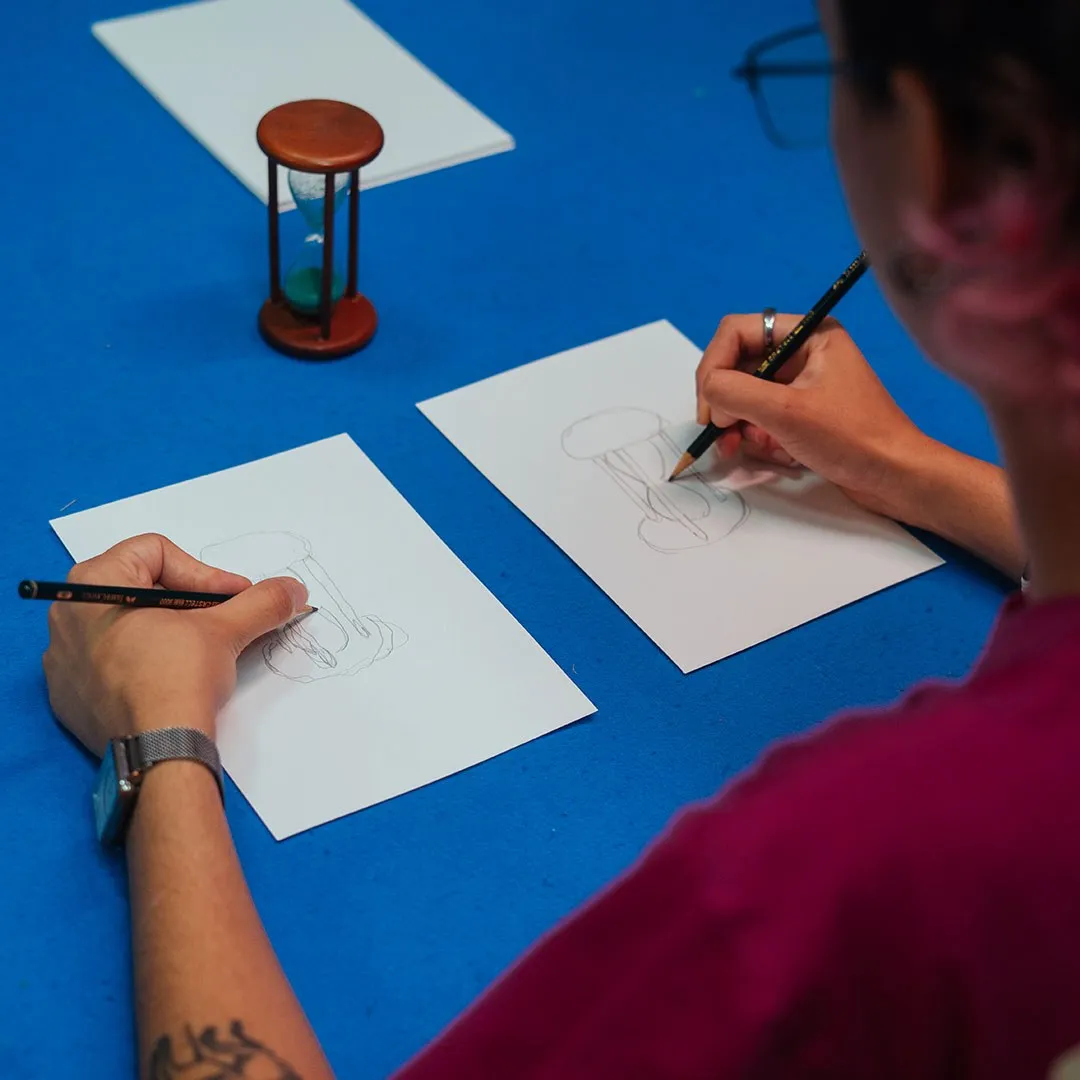 Mãos desenhando a lápis sobre folhas brancas numa mesa azul, ao lado de uma ampulheta