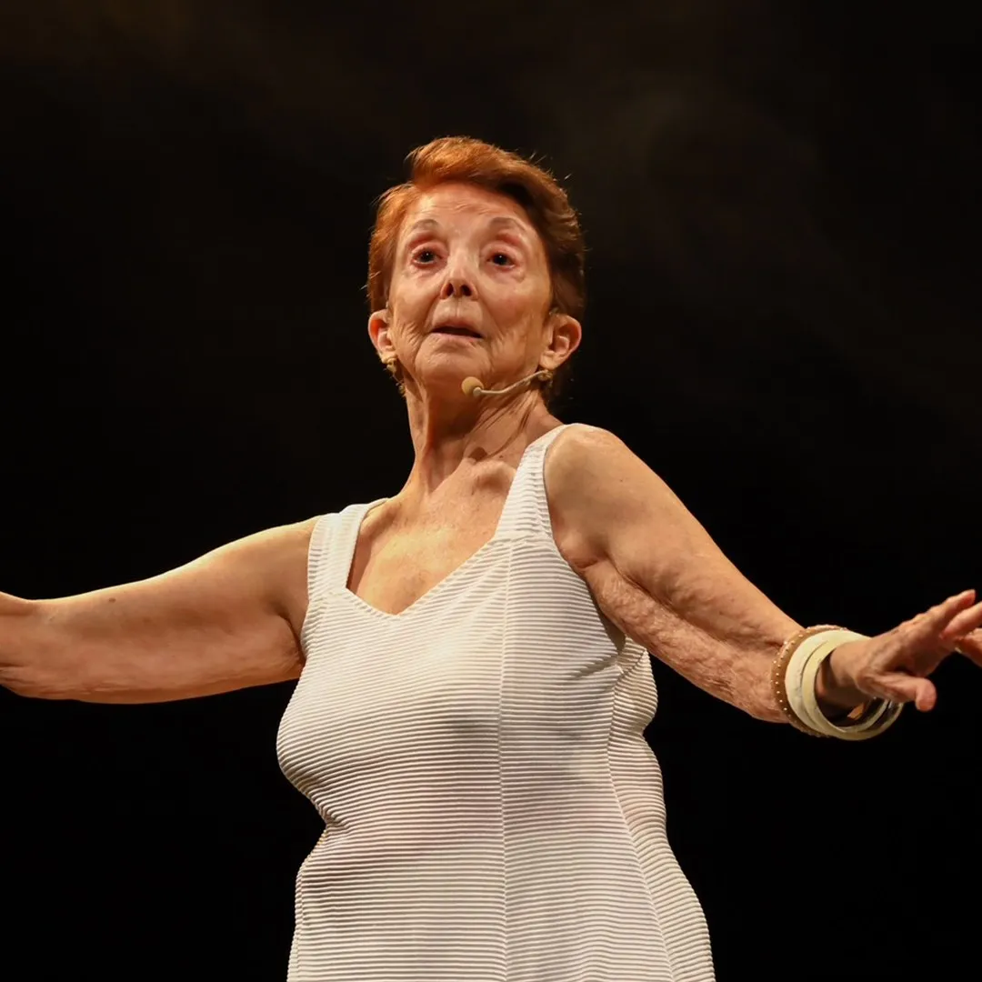 Atriz idosa de cabelo ruivo e regata branca com os braços abertos em palco escuro