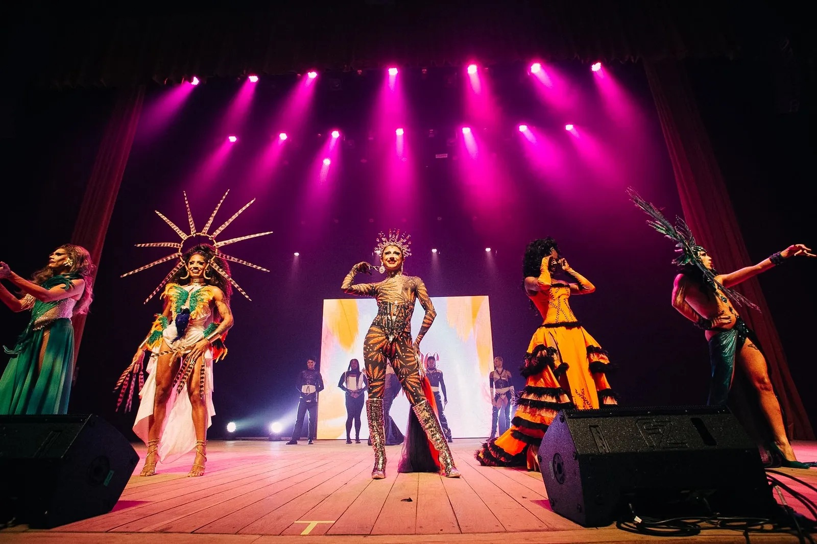 Artistas drag com figurinos coloridos e brilhantes no palco sob luzes cor-de-rosa