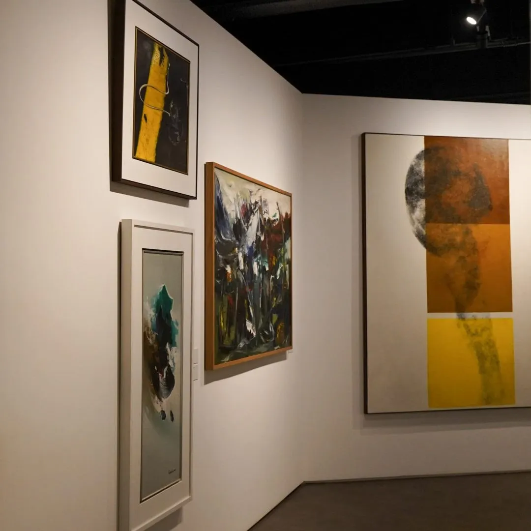 Sala expositiva com pinturas emolduradas em paredes brancas e uma tela grande em tons de ocre e amarelo